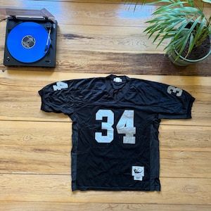 May include: Black football jersey with the number 34 in white. The jersey has white numbers on the shoulders and a small patch with text. A record player with a blue record is in the top left corner.