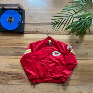 Puede incluir: Una chaqueta bomber de satén roja con rayas amarillas y blancas en las mangas y un logotipo de equipo en el pecho. El logotipo es una flecha roja con un contorno blanco y las letras "KC" en blanco.