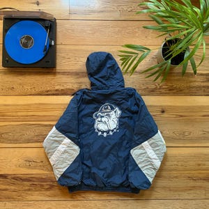 Puede incluir: Una chaqueta azul marino con capucha y detalles en blanco y gris. La chaqueta tiene un logotipo de mascota de bulldog en la espalda.