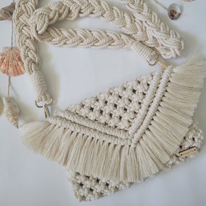 May include: Cream-colored macrame crossbody bag with a braided strap and fringe detail. The bag features a woven pattern and gold-colored hardware. Seashells are arranged around the bag.