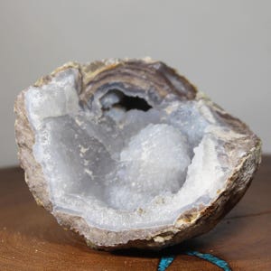 May include: A halved geode with a rough, brown exterior and a crystalline interior. The interior features a cluster of small, white crystals and a light blue hue. The geode is displayed on a wooden surface.