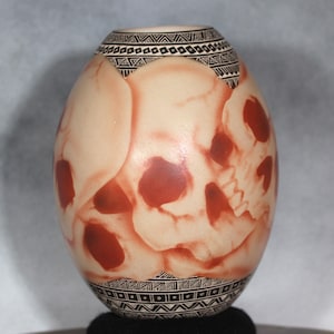 May include: An egg-shaped ceramic vase with a cream base and red skull design. The vase features black geometric patterns at the top and bottom. The skull design is a prominent feature, adding a unique and artistic touch to the decorative vase.