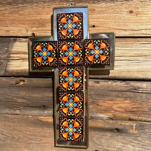 May include: A decorative cross with a mirrored background and a brass frame. The cross is adorned with colourful square tiles featuring orange, blue, and yellow floral patterns. The cross is hanging on a wooden wall.