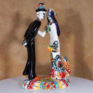 Puede incluir: Figura de cerámica del Día de Muertos que representa a una pareja de esqueletos. El hombre viste un traje negro y sombrero de copa, la mujer un vestido blanco con diseños florales y de mariposas. La base es de colores brillantes.