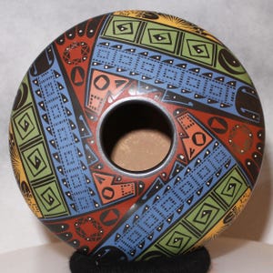 May include: A round, ceramic vessel with a central hole, featuring a vibrant geometric design. The artwork incorporates blue, green, red, and yellow patterns on a dark background. The vessel is likely a decorative piece or a functional item.