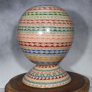 May include: A decorative, hand-painted sphere with a matching pedestal base. The sphere and base feature repeating geometric patterns in a variety of colours, including red, green, blue, and beige. The object is displayed on a wooden surface.