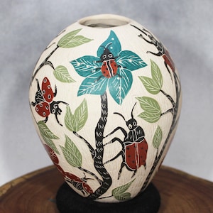 Puede incluir: Jarrón de cerámica color crema con un diseño pintado a mano de insectos, hojas y flores. Los insectos son rojos y negros, las hojas son verdes y la flor es turquesa. Un artículo decorativo para el hogar.