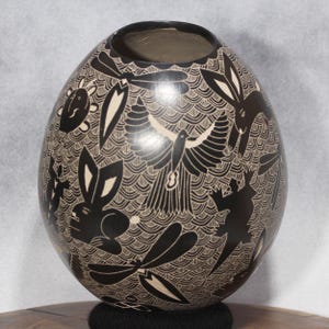 May include: A black and white ceramic vase with an egg-like shape. The vase is decorated with etched designs of animals, including rabbits, birds, and dragonflies, set against a wavy background. The rim of the vase is dark, and the interior is a lighter shade.