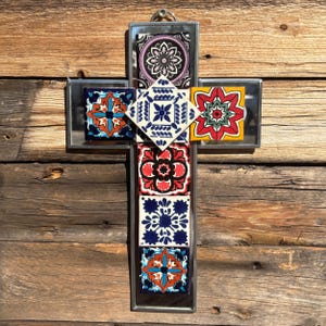 May include: A decorative cross composed of patterned ceramic tiles set within a silver-coloured frame. The tiles display floral and geometric designs in blue, red, yellow, and purple. The cross is set against a rustic wooden backdrop.