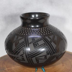Puede incluir: Un jarrón de cerámica negro con un acabado brillante. El jarrón presenta un diseño geométrico en un tono más claro de negro, con un patrón punteado cerca de la parte superior. El jarrón está sobre una superficie de madera.