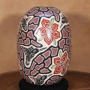 May include: A hand-painted ceramic vase with a geometric pattern in black and purple. The vase is decorated with red and white flowers and black hummingbirds.