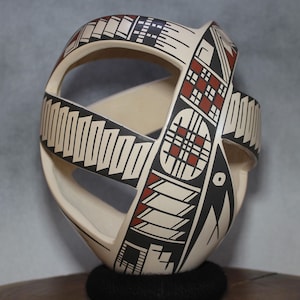 May include: A cream-colored, hand-painted ceramic vessel with an open, interwoven design. The vessel features geometric patterns in black, red, and white. The vessel is resting on a black, textured base.