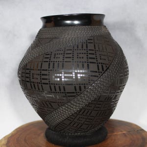 May include: A dark brown ceramic vase with a black rim and base. The vase features a geometric pattern of squares and diagonal bands with a textured, woven appearance. The vase is displayed on a wooden surface.