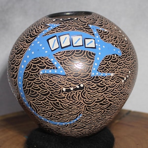 May include: A spherical ceramic vase, hand-painted with a black background and a repeating pattern of curved lines. A light blue lizard design with white dots and rectangular shapes decorates the vase. The vase has a dark brown opening.