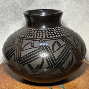Puede incluir: Un jarrón de cerámica negro con cuerpo redondeado y cuello estrecho. El jarrón presenta un diseño geométrico en un tono más claro de negro, con una banda de pequeños triángulos cerca de la parte superior. El jarrón se exhibe sobre una superficie de madera.