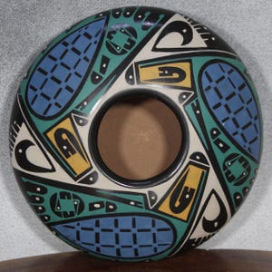 May include: A round, ceramic vessel with a central opening. The exterior is decorated with geometric patterns in black, blue, green, and yellow against a white background. The design features stylized shapes and lines.