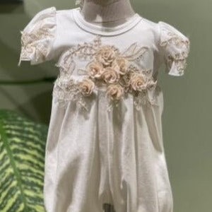 May include: A white baby romper with lace trim and a floral design. The romper has short sleeves and a gathered skirt. The floral design is made of beige fabric flowers.
