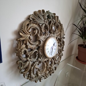 Puede incluir: Reloj de pared redondo y ornamentado con esfera dorada y números romanos. El reloj está enmarcado en un marco decorativo, con remolinos y motivos florales, en un tono dorado apagado. El reloj está montado en una pared de color claro.