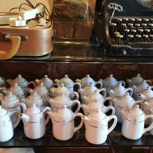 Könnte beinhalten: Eine Sammlung von 20 weißen Keramik-Teekannen mit blauen Verzierungen und einem schwarzen Buchstaben 'S' auf jeder. Die Teekannen sind in Reihen auf einem Glasregal angeordnet.