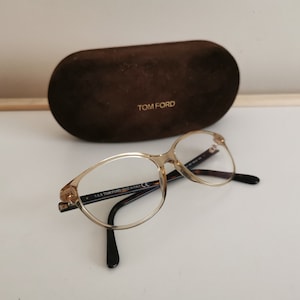Może przedstawiać: Okulary w złotej oprawie z przezroczystymi szkłami i czarnymi zausznikami. Okulary leżą na białym tle obok brązowego etui z nadrukiem "TOM FORD".
