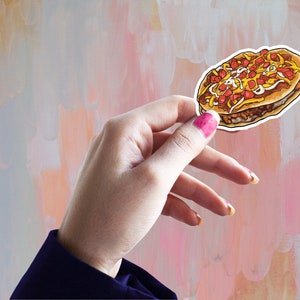 May include: A colorful sticker featuring a cartoon illustration of a loaded sandwich. The sandwich has layers of meat, cheese, and toppings. The sticker is held against a soft, abstract background with pink, peach, and white brushstrokes.