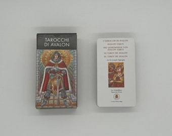 May include: Two tarot card decks, one with a black background and the text "TAROCCHI DI AVALON" in white, and the other with a white background and the text "TAROCCHI DE AVALON AVALON TAROT DIE GEHEIMNISSE VON AVALON TAROT LE TAROT DE AVALON EL TAROT DE AVALON Art by Joseph Viglioglie" in black. The card on the left shows a man in a red and gold robe holding a sword. The card on the right shows a man and a woman in a forest setting.