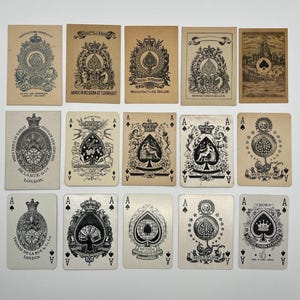 Peut inclure: Un ensemble de cartes à jouer anciennes, avec des motifs d'As de Pique ornés. Les cartes présentent des illustrations complexes, des armoiries et du texte, dont "Thomas De La Rue & Co London" et "Made in Belgium at Turnhout". Les cartes sont disposées sur une surface blanche.