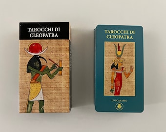 May include: Two tarot card decks with a blue and green background. The front of the deck on the left shows a green figure with a red sun and a black background. The front of the deck on the right shows a woman in a red dress with a gold headpiece and a blue background. The text on both decks reads "Taroocchi di Cleopatra".