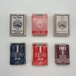 Peut inclure: Six jeux de cartes disposés sur deux rangées. La rangée supérieure comprend un jeu blanc 'NAIPE ESPAÑOL', un jeu 'TOURISM-202' à rayures rouges et un jeu blanc '202 TURISMO'. La rangée inférieure contient un jeu bleu 'TURISMO 101', un jeu rouge 'TURISMO 101' et un jeu bordeaux 'NAIPE ESPAÑOL 121'.