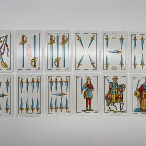 May include: A set of ten playing cards from a Spanish deck, featuring swords. The cards are numbered 1 through 10, with the 10 card showing a king in a red and gold robe. The cards are on a white background.