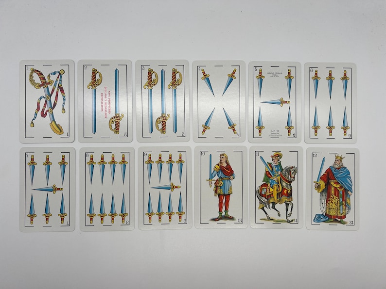 May include: A set of ten playing cards from a Spanish deck, featuring swords. The cards are numbered 1 through 10, with the 10 card showing a king in a red and gold robe. The cards are on a white background.