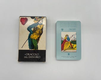 May include: A deck of Oracolo del Danubio oracle cards in a rectangular box featuring a person in a yellow and green outfit with a red heart. Beside it, a single light blue card displays a seated figure in yellow and red robes with text.