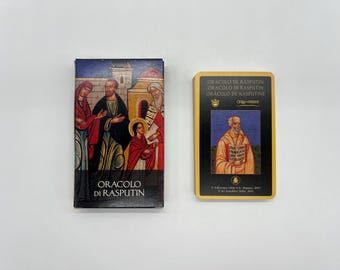 May include: A deck of Oracolo di Rasputin oracle cards featuring a rectangular box with a religious-style illustration of figures in robes and a single card with a black border, gold trim, and a central portrait of a bearded man holding a book.