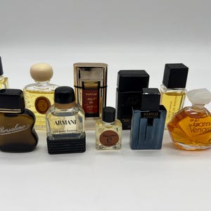 May include: A selection of miniature perfume bottles in various shapes and sizes. The bottles feature different colours, including clear, brown, black, and gold. Some bottles have black caps, while others have gold or white tops. The bottles are arranged on a white surface.