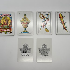 May include: A set of playing cards featuring a variety of designs, including a woman's portrait, a vase, a sword, and a vegetable. The cards are white with black borders and feature the text "Victoria" and "Fournier".