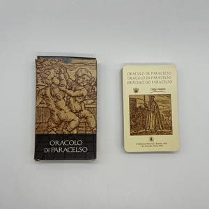 Puede incluir: Un mazo de cartas de oráculo y su caja. La caja presenta una ilustración marrón de querubines. Las cartas son de color crema con texto y un dibujo de una figura vestida con una túnica. El texto de la caja dice "ORACULO DI PARACELSO".
