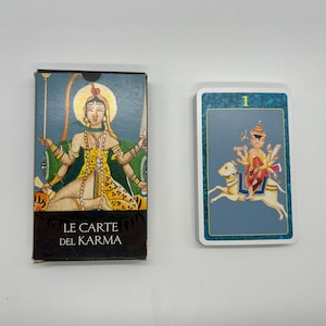 May include: A tarot card deck and a single card. The deck box is black with the text "LE CARTE DEL KARMA" and an illustration of a deity. The card shows a figure riding a camel.
