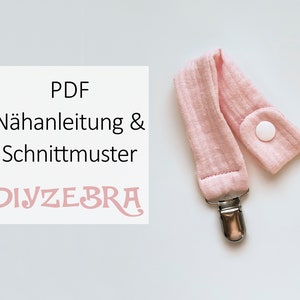 May include: A pink fabric baby teether with a white button and a silver clip. The text "PDF Nähnanleitung & Schnittmuster DIYZEBRA" is on a white background.