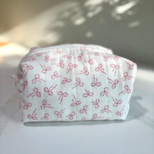 May include: A white quilted cosmetic bag with a repeating pattern of pink bows. The bag is rectangular with a zipper closure. The background is a light color.