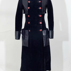 May include: A black coat with a double-breasted design, featuring red buttons and leather accents on the collar, cuffs, and pockets. The coat is displayed on a mannequin, showcasing its tailored fit and vintage style.