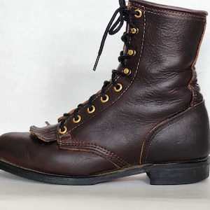 May include: A brown leather work boot with a lace-up design and a rounded toe. The boot features a dark brown color, gold-colored eyelets, and a fringe detail on the toe. The sole is black.