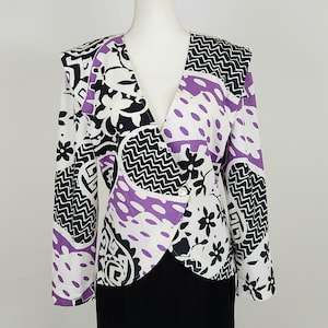May include: A white, black, and purple patterned blazer with a V-neck and long sleeves. The blazer features a mix of floral, polka dot, and geometric designs. The blazer is buttoned and worn over a black garment.
