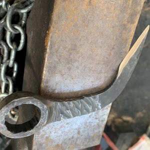 May include: A forged metal hook with a wrench head on one end. The hook is curved and has a textured surface. The hook is resting on a metal surface.