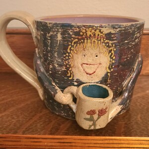 May include: A ceramic mug with a dark blue and purple textured exterior featuring a sculpted face with yellow hair. The mug has a large handle and a smaller, attached miniature mug held by sculpted arms, decorated with two small red flowers.