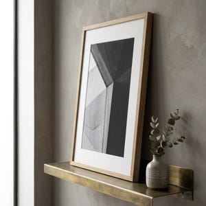 May include: A framed monochrome architectural print in a light wood frame on a brass shelf. A small ceramic vase with greenery is on the shelf, against a neutral wall.