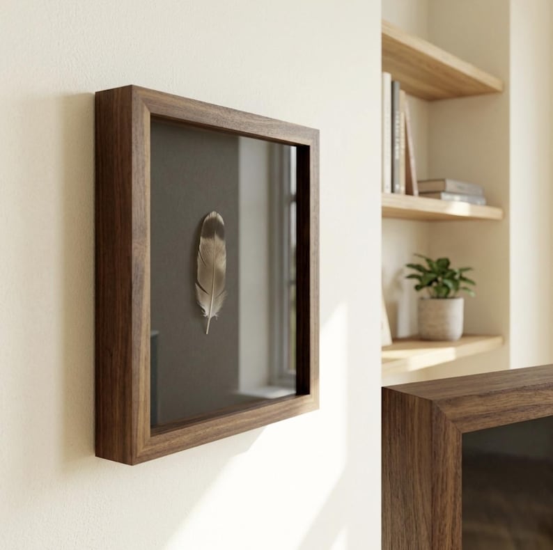 May include: A framed feather display. The feather is a light brown and gray color, set against a dark gray background. The frame is made of dark brown wood and is mounted on a white wall. The display is a decorative piece.