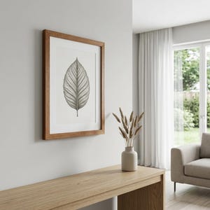 May include: A framed botanical print depicting a detailed leaf illustration in grey and white tones. The artwork is set within a warm wooden frame, mounted on a light grey wall. A minimalist piece of home decor, suitable for various interior styles.