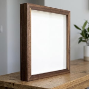 May include: A square, dark brown wooden picture frame with a white interior, standing on a wooden table. The frame has a simple, modern design and is empty, ready for a photo or artwork. The frame is approximately 12 inches by 12 inches.