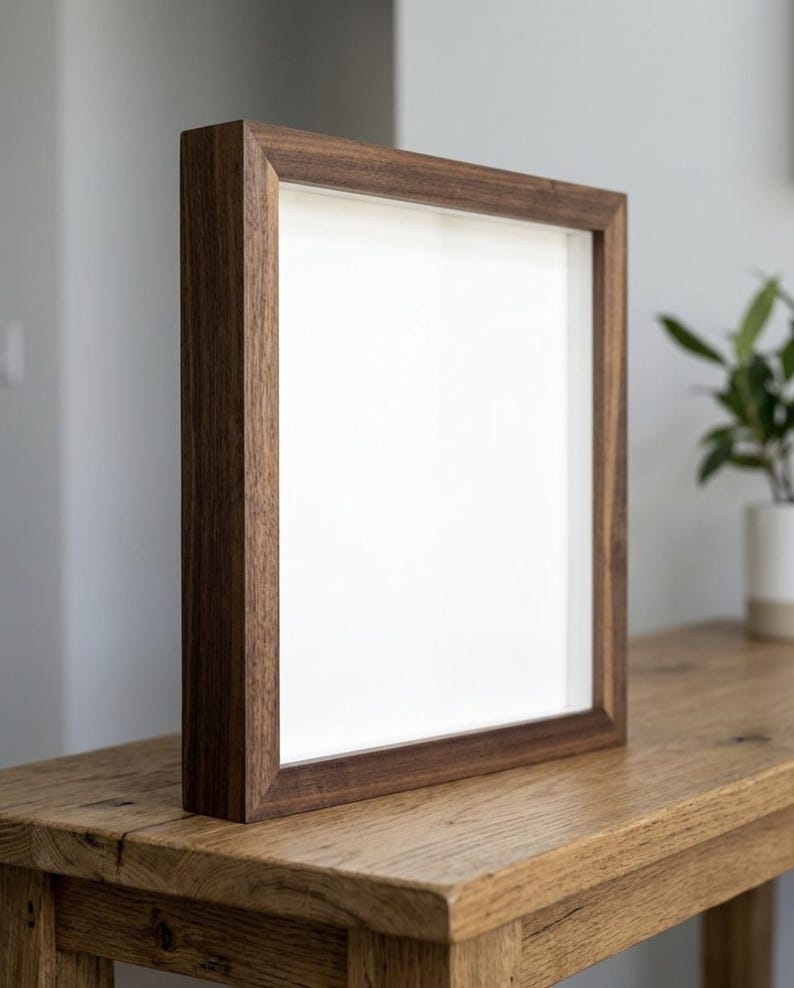 May include: A square, dark brown wooden picture frame with a white interior, standing on a wooden table. The frame has a simple, modern design and is empty, ready for a photo or artwork. The frame is approximately 12 inches by 12 inches.