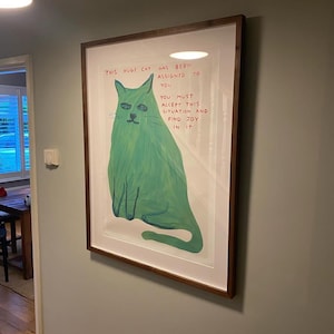 May include: A framed art print featuring a large green cat illustration. The artwork includes text that reads "THIS HUGE CAT HAS BEEN ASSIGNED TO YOU. YOU MUST ACCEPT THIS SITUATION AND FIND JOY IN IT."